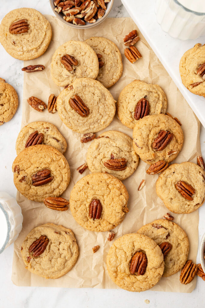 Butter pecan cookies, stacked up against each other on a piece of parchment paper with extra pecans on the table.