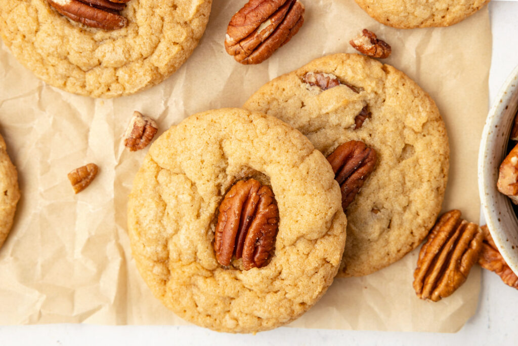 Butter pecan cookies, stacked up against each other on a piece of parchment paper with extra pecans on the table.