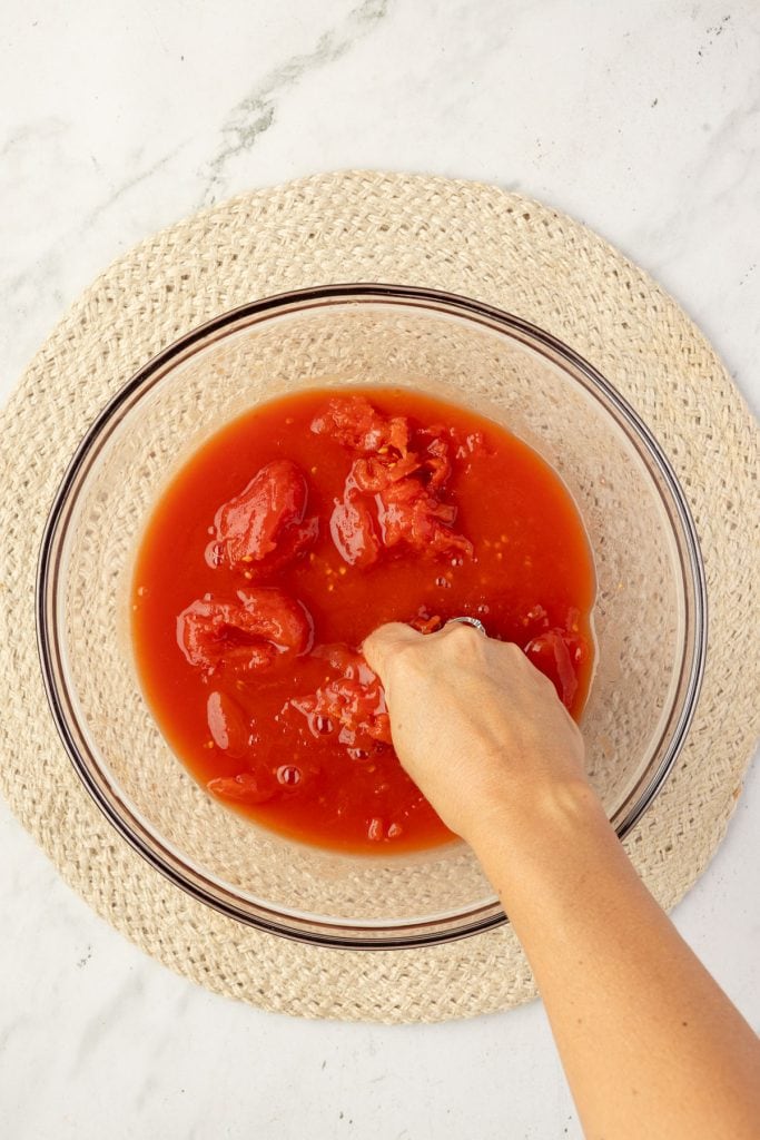 Hand crushing tomatoes in a bowl.