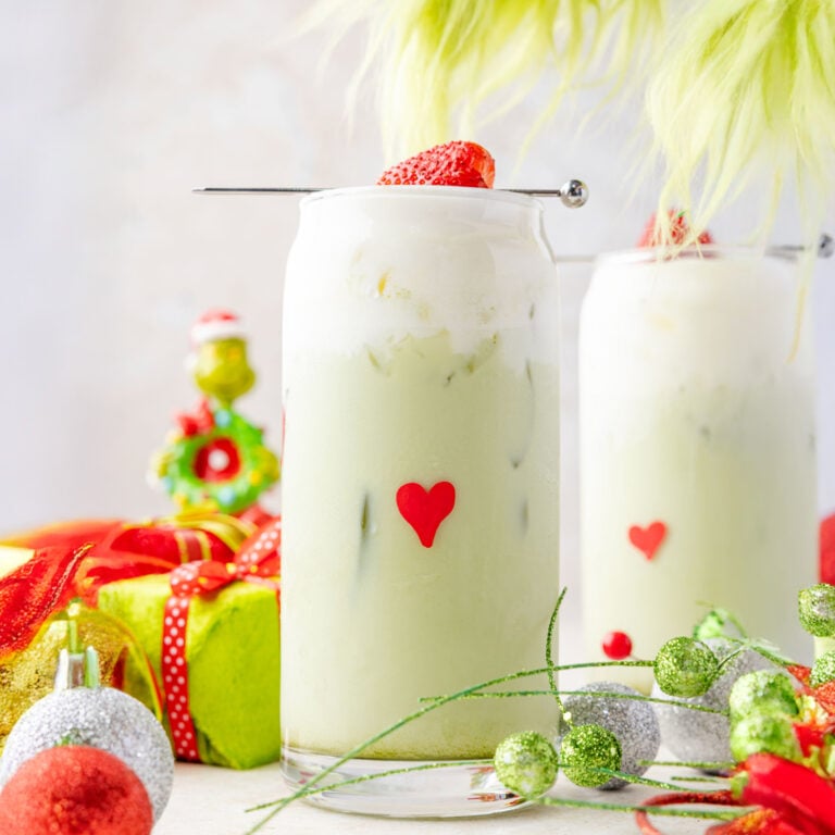 Christmas scene with Grinch decorations and two iced grinch Matcha lattes with the Grinch hand coming into steal a latte.