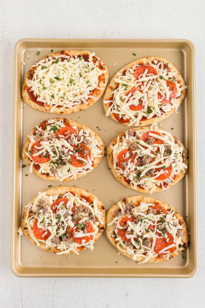 raw naan flatbread pizza on a sheet pan topped with cheese, pepperoni, and sausage.
