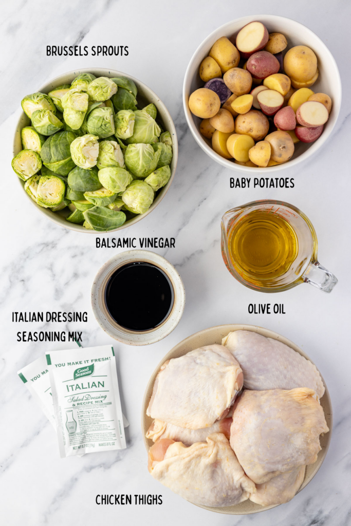 Potatoes, brussels sprouts, oil, balsamic vinegar, Italian seasoning, packets, and chicken thighs in individual bowls on a marble table.