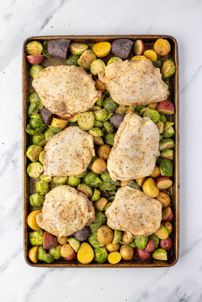 Raw seasoned chicken thighs on top of brussels sprouts and potatoes on a sheet pan.
