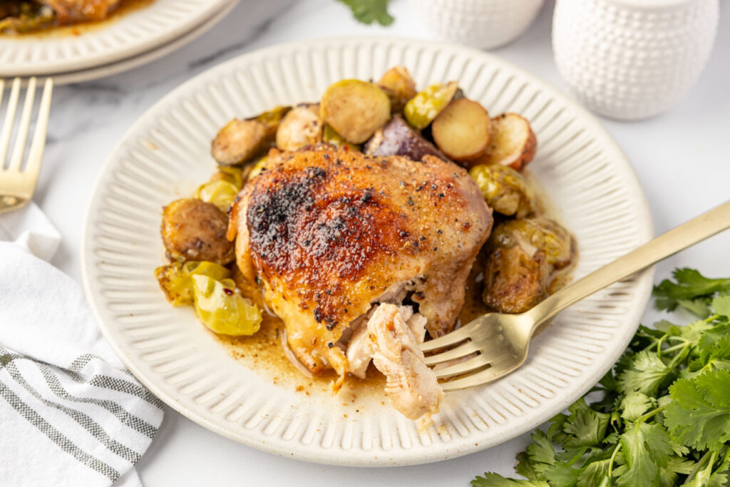 Baked chicken thigh with roasted brussels sprouts, and potatoes on a plate with a fork holding a piece of the chicken thigh cut open.