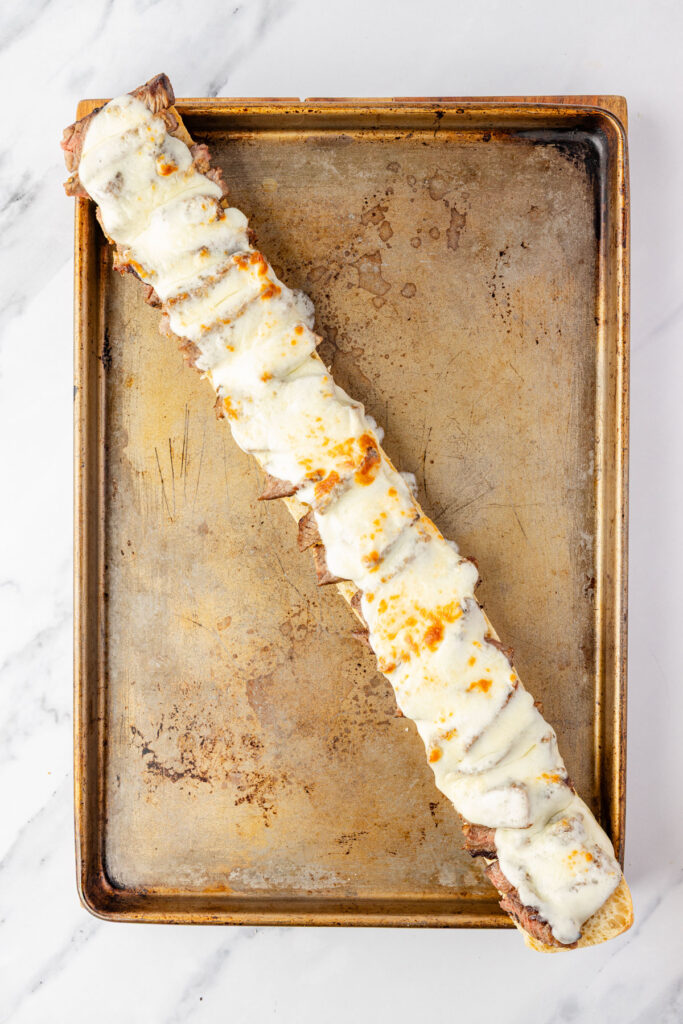 Bottom half of steak sandwich on a sheet pan after coming out of the oven with melted cheese