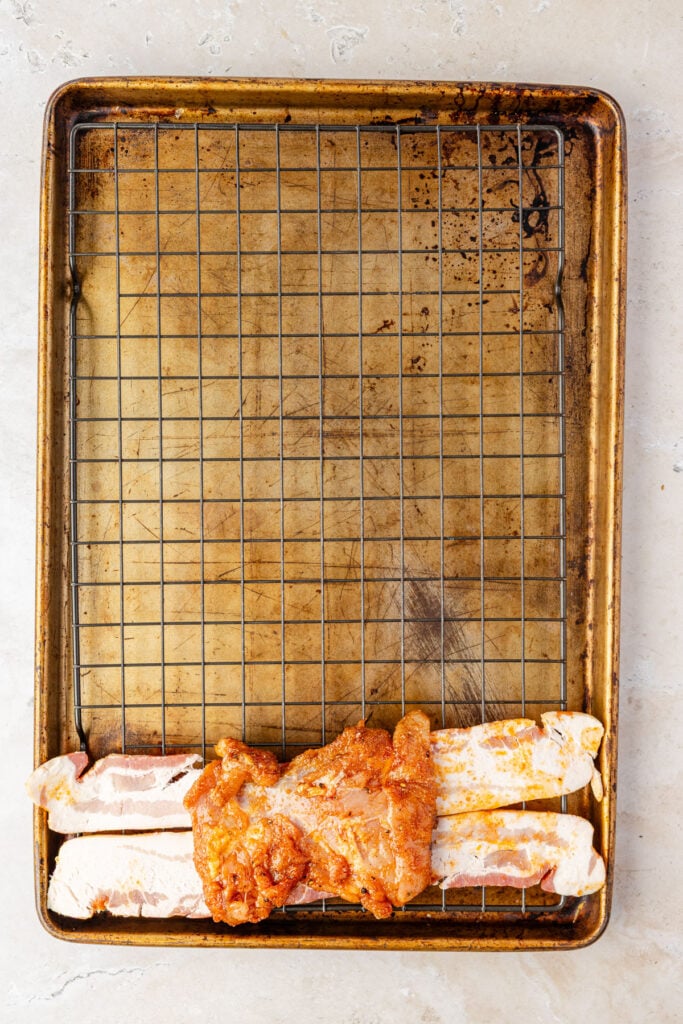 Seasoned chicken on top of two strips of bacon on a sheet pan