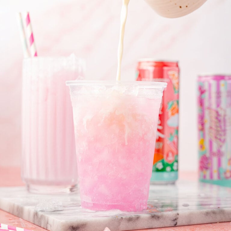 Heavy cream being poured into a cup of Alani on a pink background with a marble slab