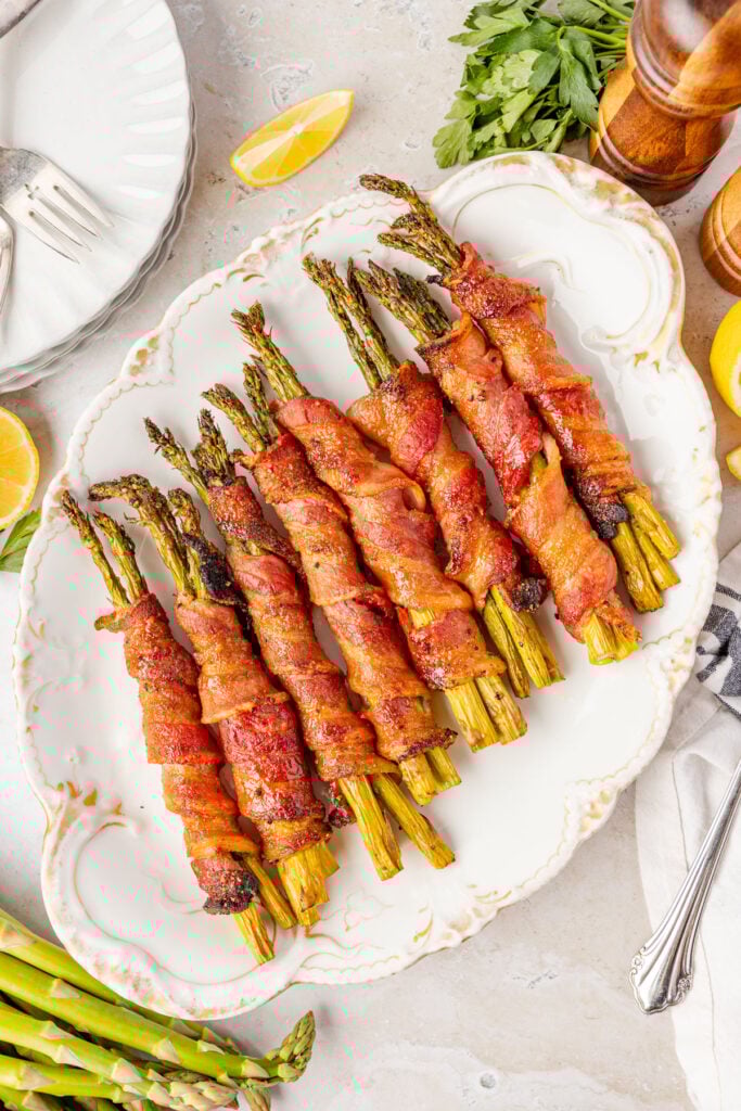 A plate of bacon wrapped asparagus with parsley and lemons around it