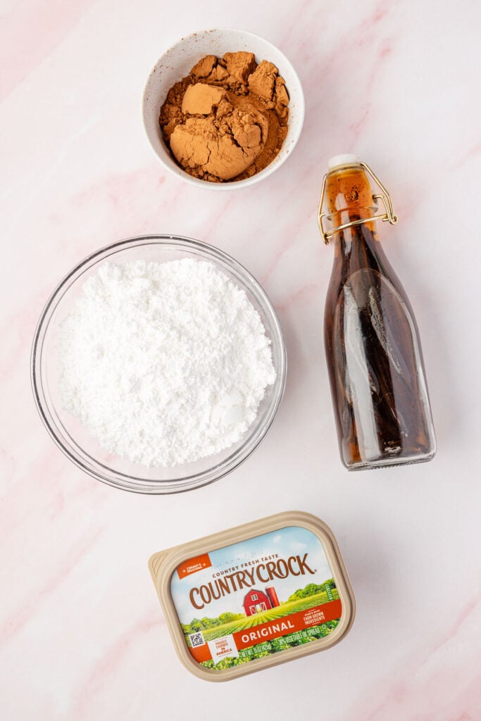 Vanilla extract in a bottle, country cock in a tub, powdered sugar in a bowl, cocoa powder in a bowl to make homemade chocolate frosting