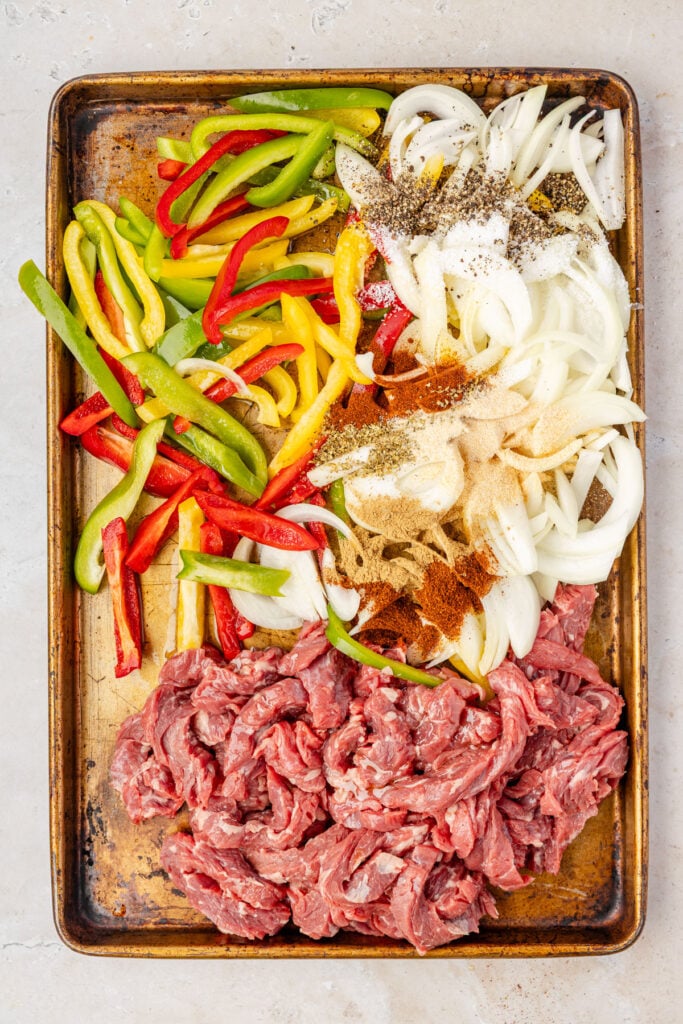 Raw seasoned steak, peppers, and onions on a sheet pan for
