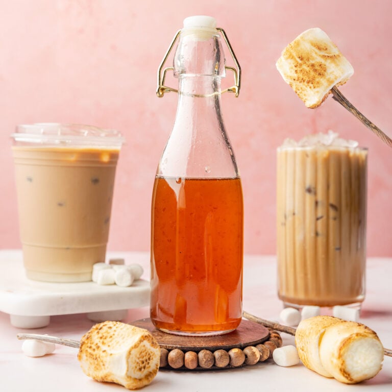 Toasted marshmallow syrup in a bottle with two iced coffees in the background with toasted marshmallows on sticks around it