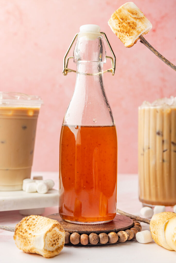 Toasted marshmallow syrup in a bottle with two iced coffees in the background with toasted marshmallows on sticks around it