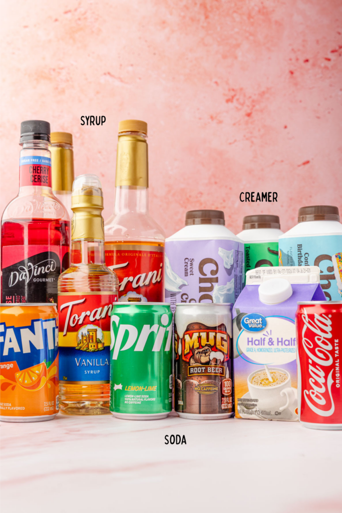 Different flavored syrups, different sodas, and different types of coffee creamer on a marble board with a pink background