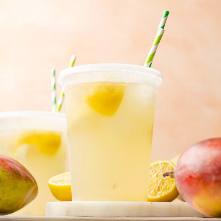 Mango Lemonade in a container with a straw and mangoes and lemon surrounding it