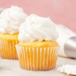 Two vanilla cupcakes with butter frosting, surrounded by cake, pans and piping bags filled with American buttercream frosting