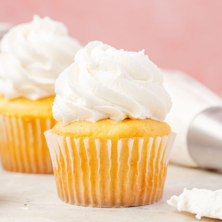 Two vanilla cupcakes with butter frosting, surrounded by cake, pans and piping bags filled with American buttercream frosting