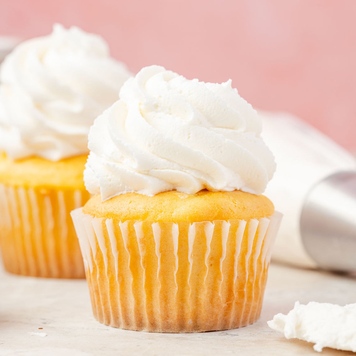 Two vanilla cupcakes with butter frosting, surrounded by cake, pans and piping bags filled with American buttercream frosting