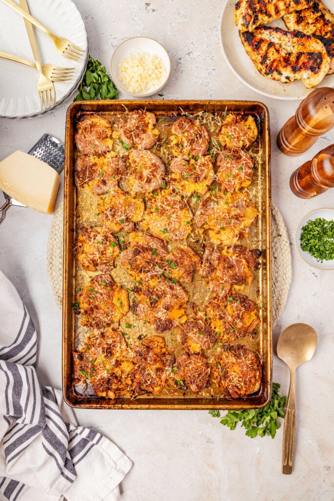 Mashed potatoes on a sheet pan with Parmesan cheese, surrounded by plates and salt and pepper and spoons and Parmesan cheese.