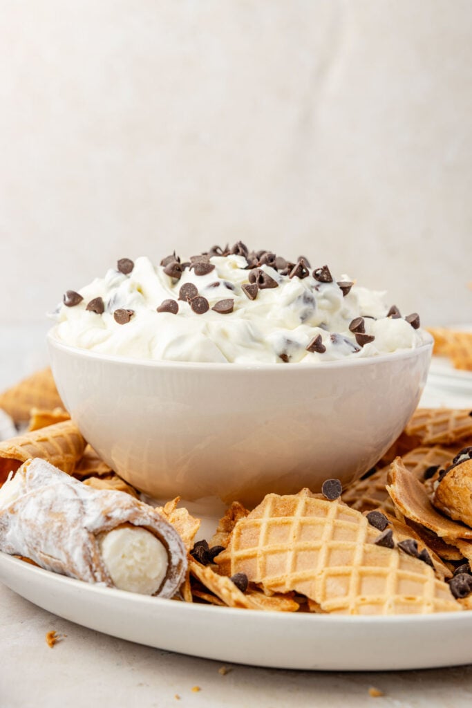 Cannoli Dip in a bowl with waffle cones and cannoli on a plate around it.