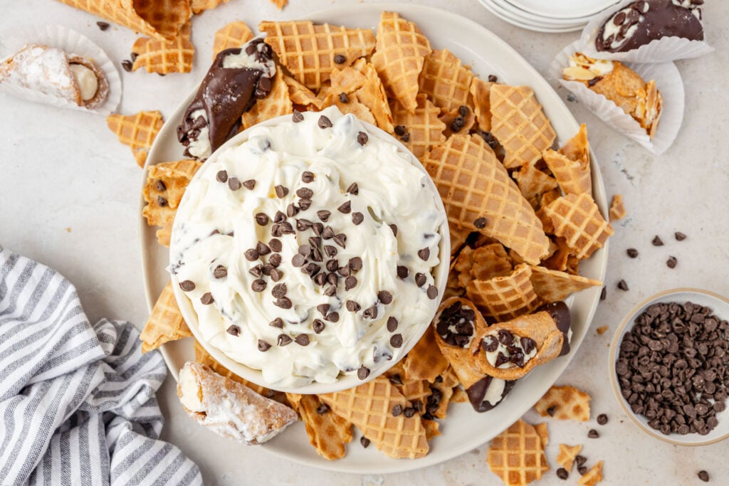 Cannoli Dip in a bowl with waffle cones and cannoli on a plate around it.