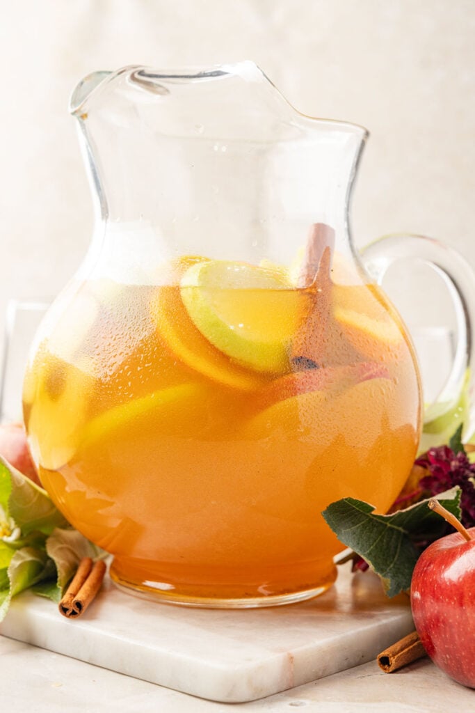 Apple Cider Sangria in a pitcher with fruit, flowers and leaves around it.