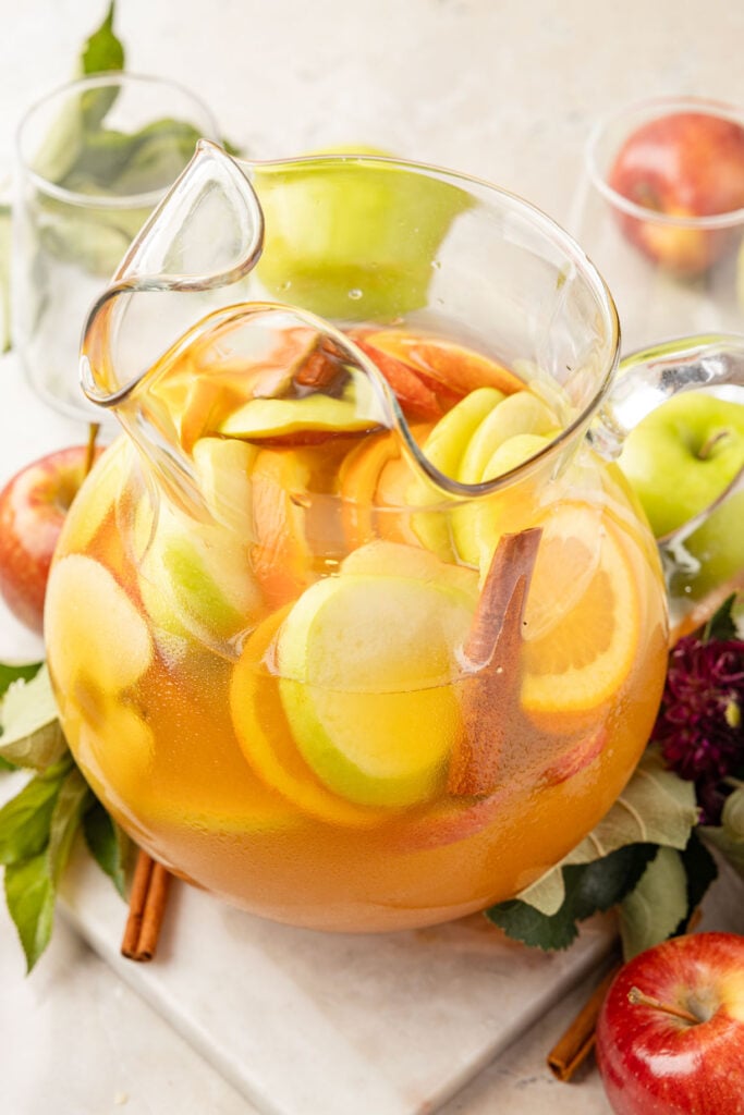 Apple Cider Sangria in a pitcher with fruit, flowers and leaves around it.
