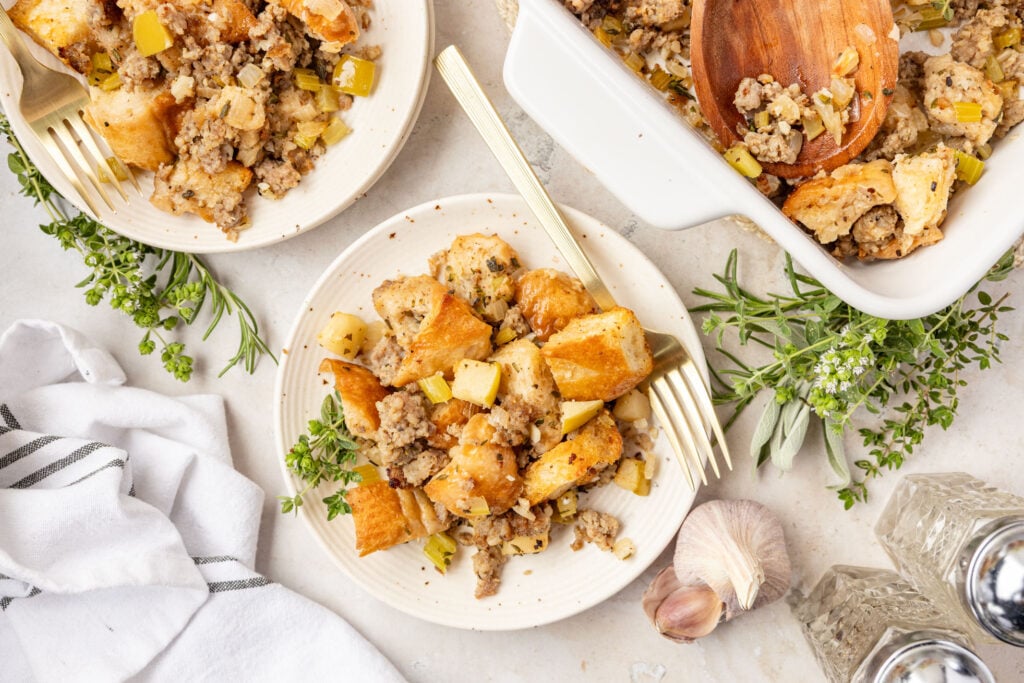 Applesauce, stuffing on a plate surrounded by herbs and garlic with the casserole pan and another plate of stuffing.