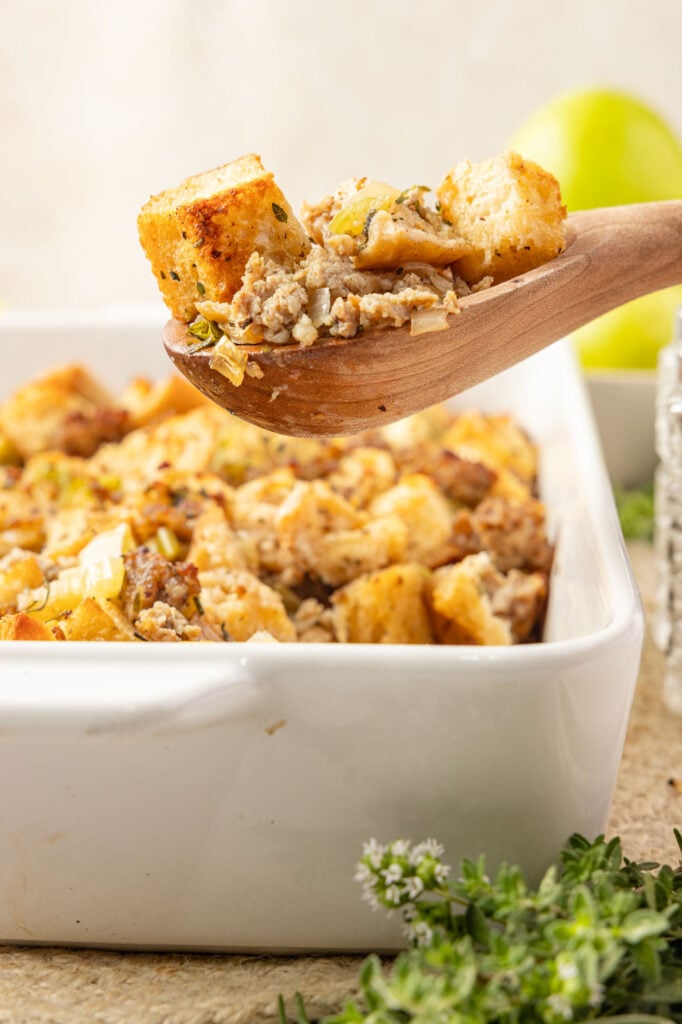 A casserole pan filled with apple sausage stuffing scooping out a big spoonful of it with a wooden spoon.
