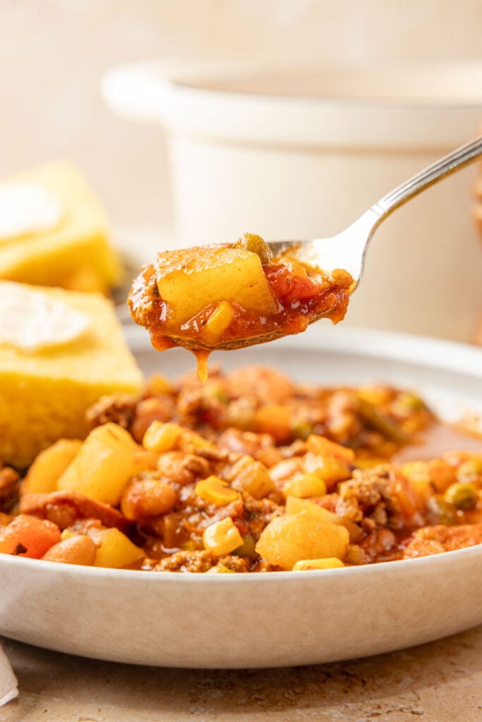 Cowboy steel in a bowl with a pot in the background and cornbread with a spoon, picking up a spoonful of the stew.