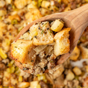 A casserole pan filled with apple sausage stuffing scooping out a big spoonful of it with a wooden spoon.