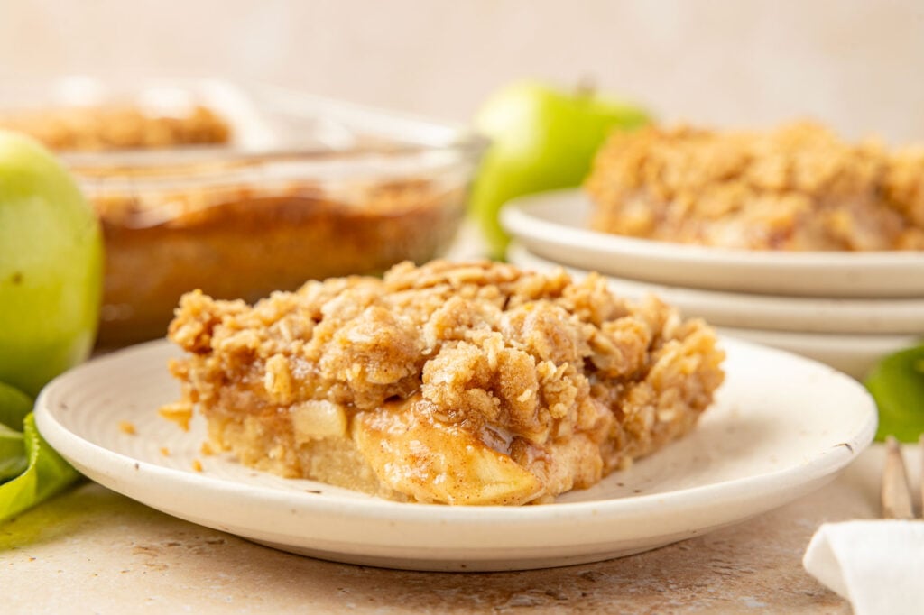 Apple pie bar on a plate with the pan of them in the background and a stack of plates with another bar and apple.