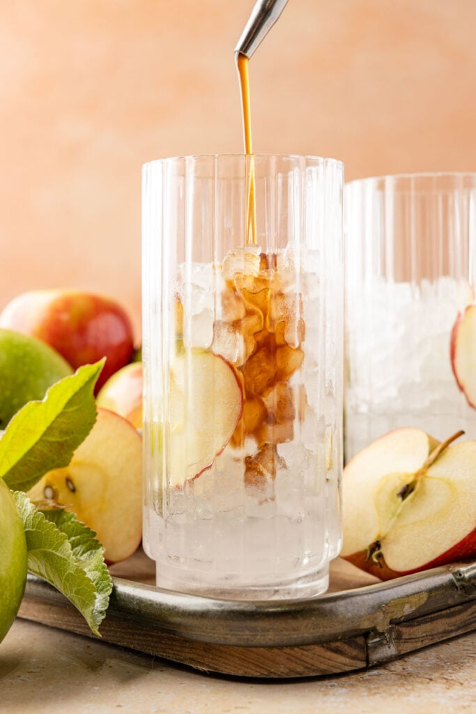 Pouring apple brown sugar syrup into a glass with an apple slice inside the glass surrounded by lots of apples and leaves.