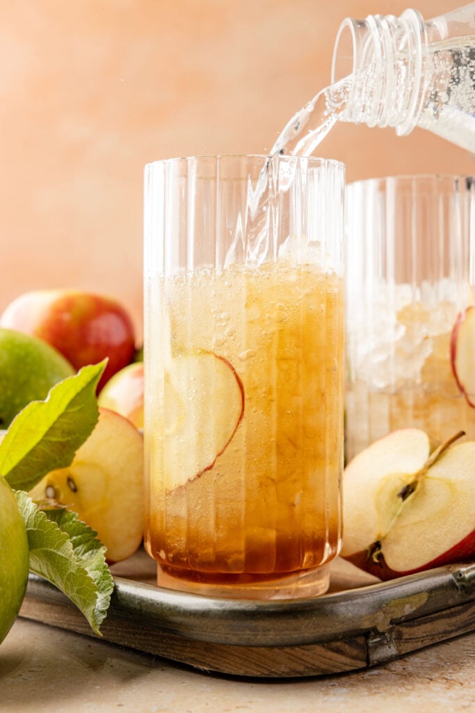 Pouring sparkling water into a glass with an apple slice inside the glass surrounded by lots of apples and leaves