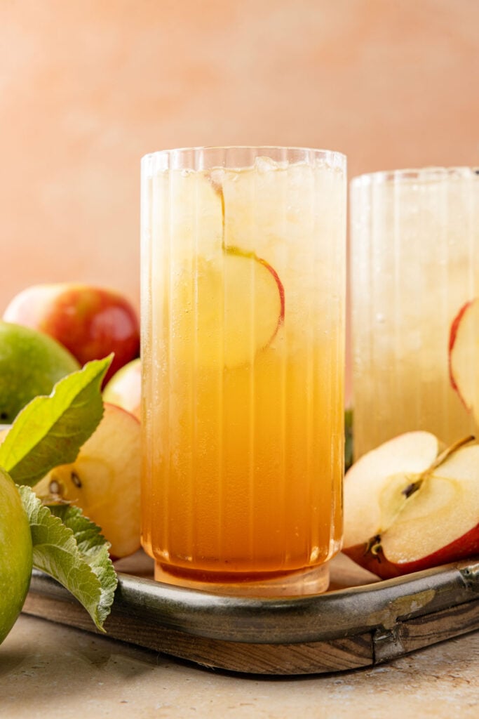 Finished Apple soda on a tray with an apple slice inside the glass surrounded by lots of apples and leaves