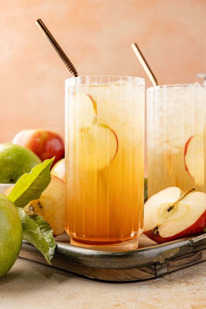 Apple soda on a tray with an apple slice inside the glass surrounded by lots of apples and leaves