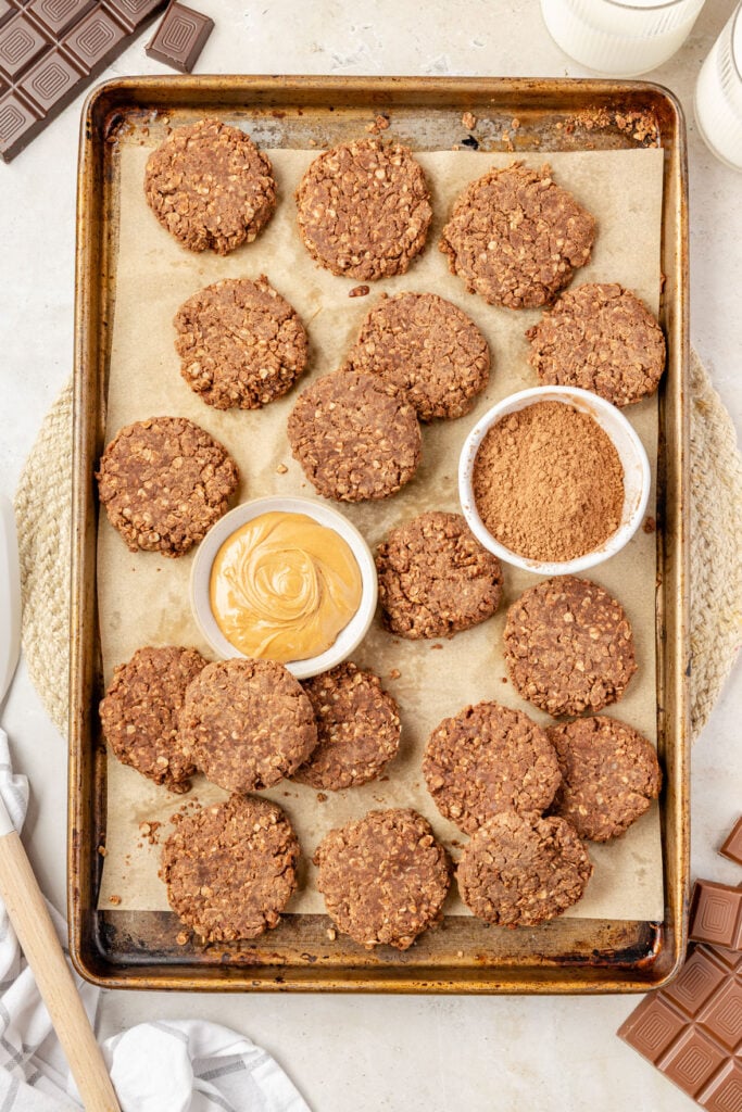 Chocolate no bake cookies on a sheet pan with a bowl of peanut butter and a bowl of cocoa powder