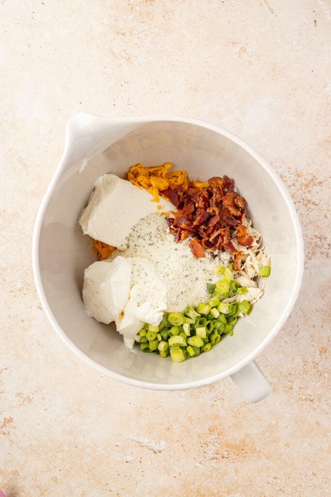 Mixing bowl full of bacon, sour cream, chicken, green onions, ranch seasoning, and cheese