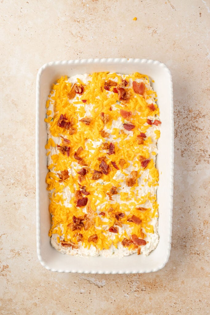 Uncooked cracked chicken dip, topped with cheese and bacon before going in the oven.