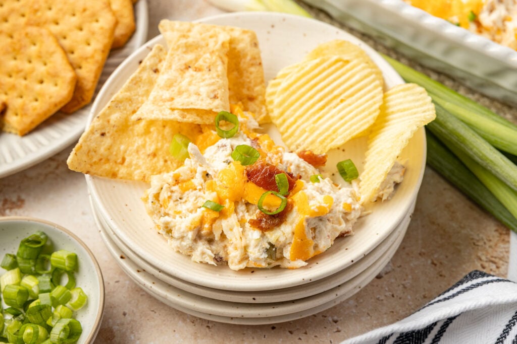 Crack chicken dip on a plate with tortilla chips and wavy potato chips, surrounded by green onions and celery and a plate of chips.
