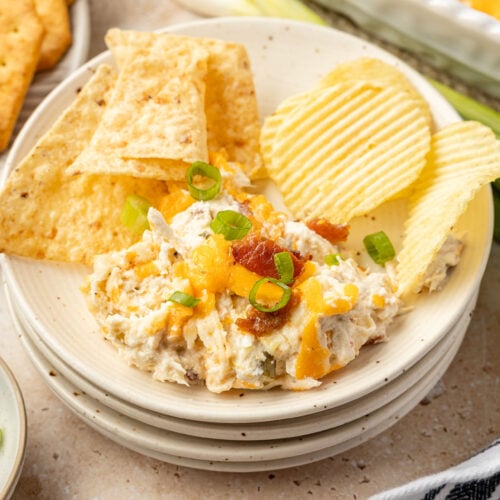 Crack chicken dip on a plate with tortilla chips and wavy potato chips, surrounded by green onions and celery and a plate of chips.