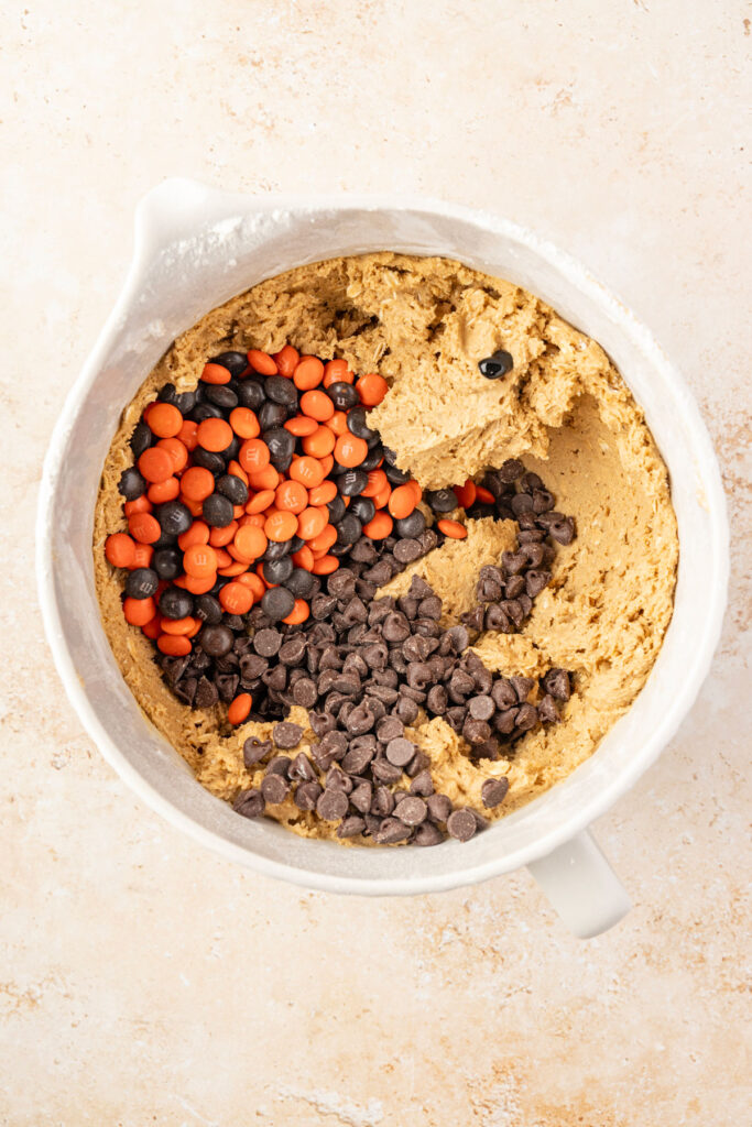 Monster cookie batter with M&Ms, chocolate chips, and purple food coloring in a mixing bowl about to be mixed.