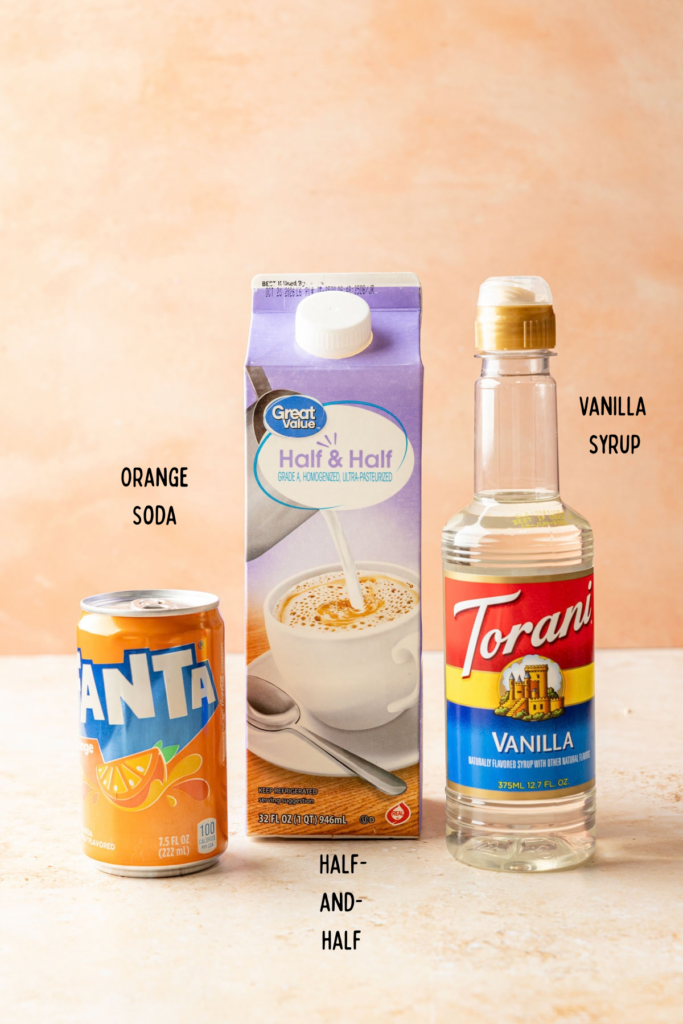 Orange soda can, carton of half-and-half, and container of vanilla syrup on a stone background.