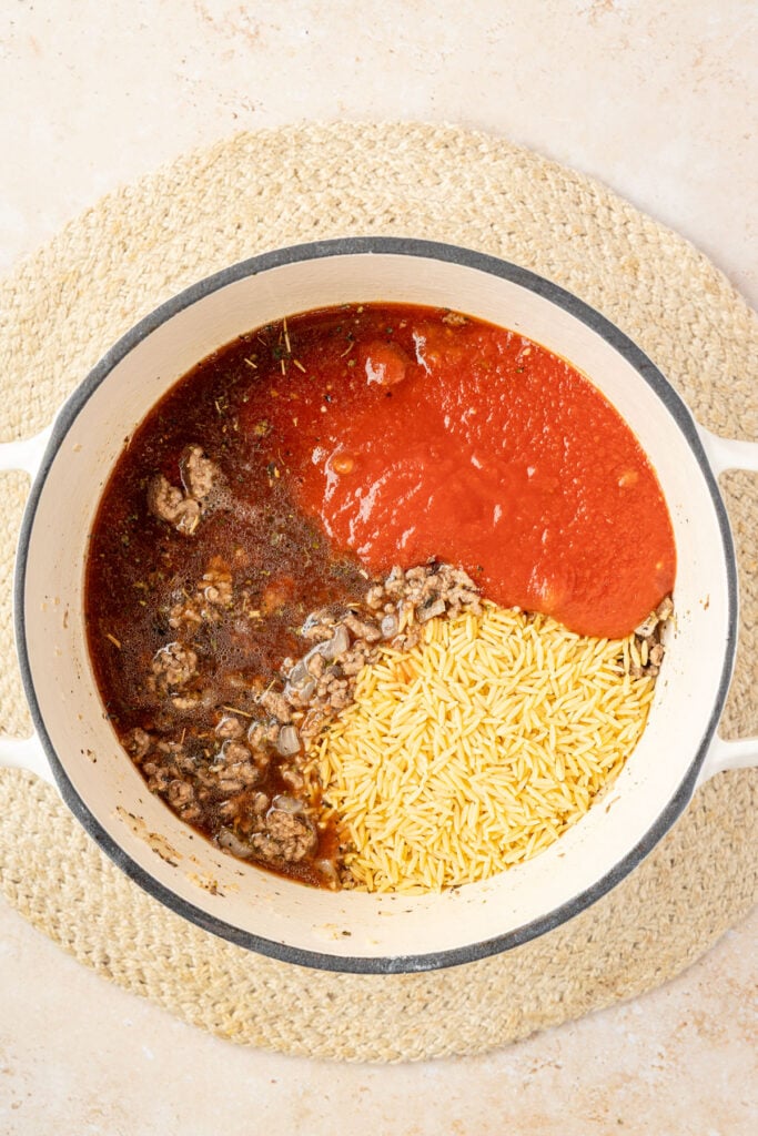 Cooked ground beef with beef broth, tomatoes, sauce, and orzo in a white Dutch oven.