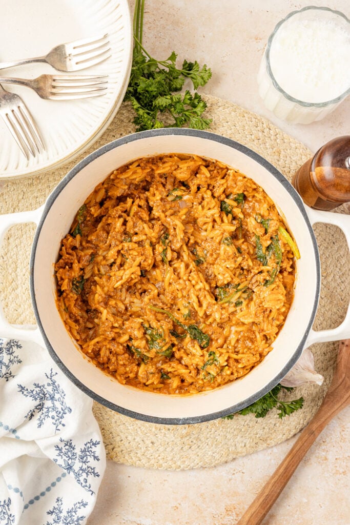 One Pot Beef Orzo in a Dutch oven on a placemat with plates and fresh herbs around it.