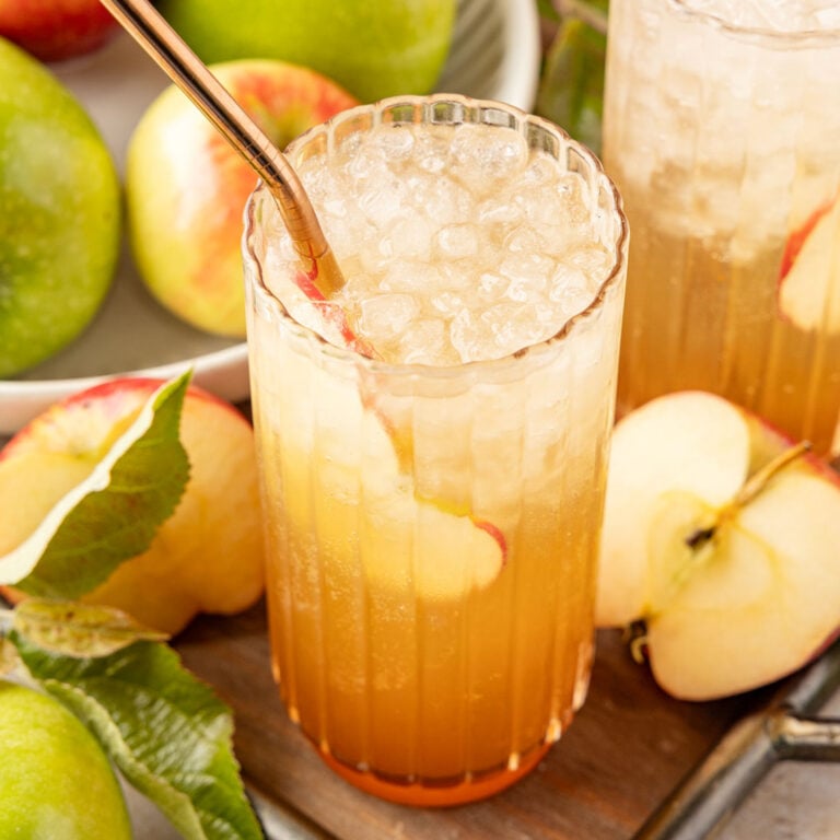 Apple soda on a tray with an apple slice inside the glass surrounded by lots of apples and leaves
