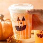 Jack-O’-Lantern Dirty Soda in a cup with a hand pick up. with a tray with pumpkins.