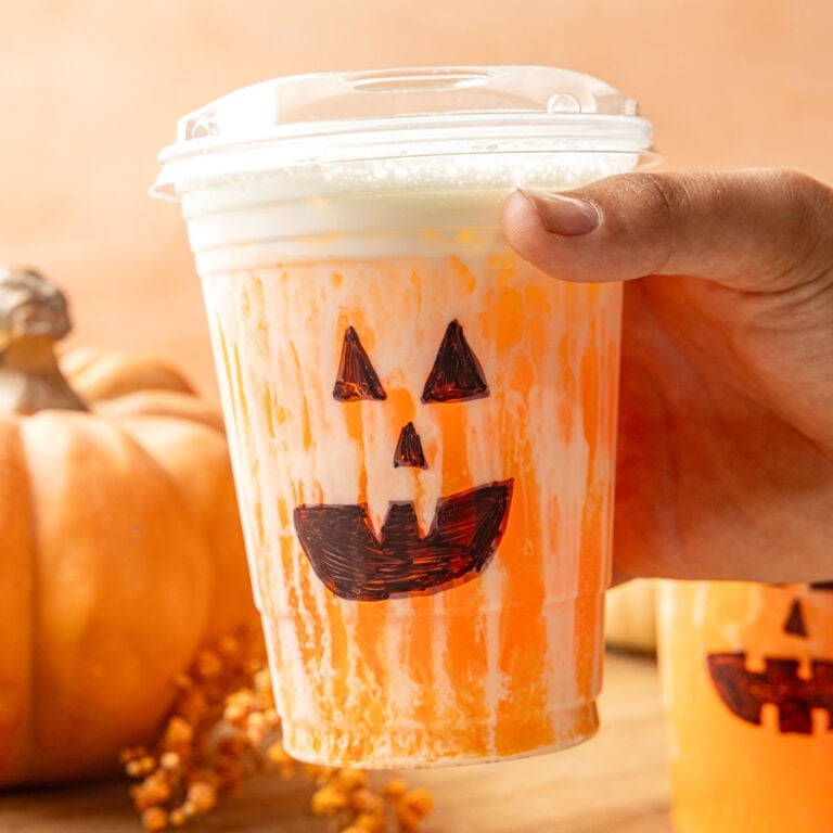 Jack-O’-Lantern Dirty Soda in a cup with a hand pick up. with a tray with pumpkins.