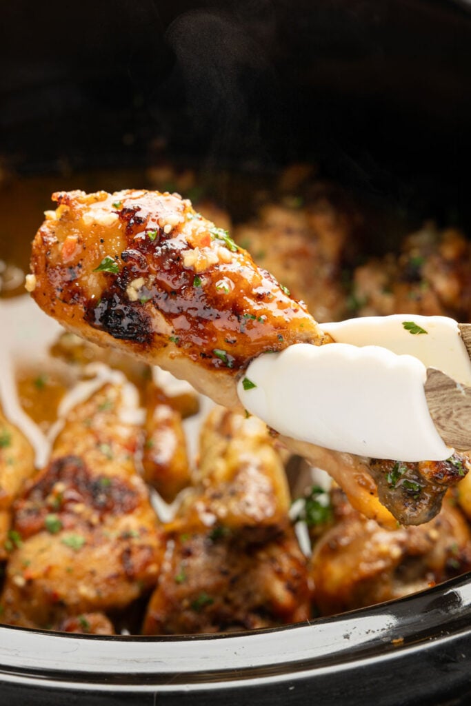 Tongues holding a piece of slow cooker, chicken drumsticks above the slow cooker.