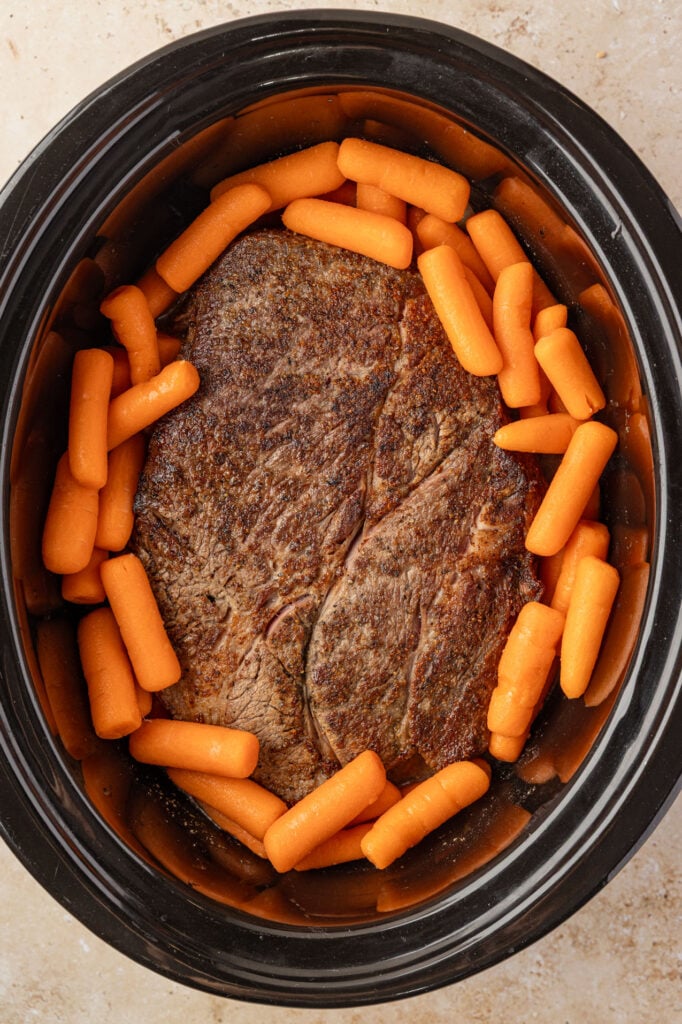 Sear chuck roast with baby carrots surrounding it in a slow cooker.