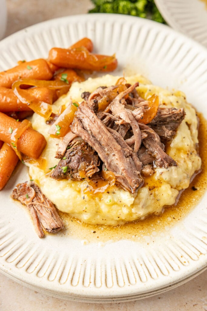 Plated slow cooker chuck roast on top of mashed potatoes with the gravy and baby carrots.